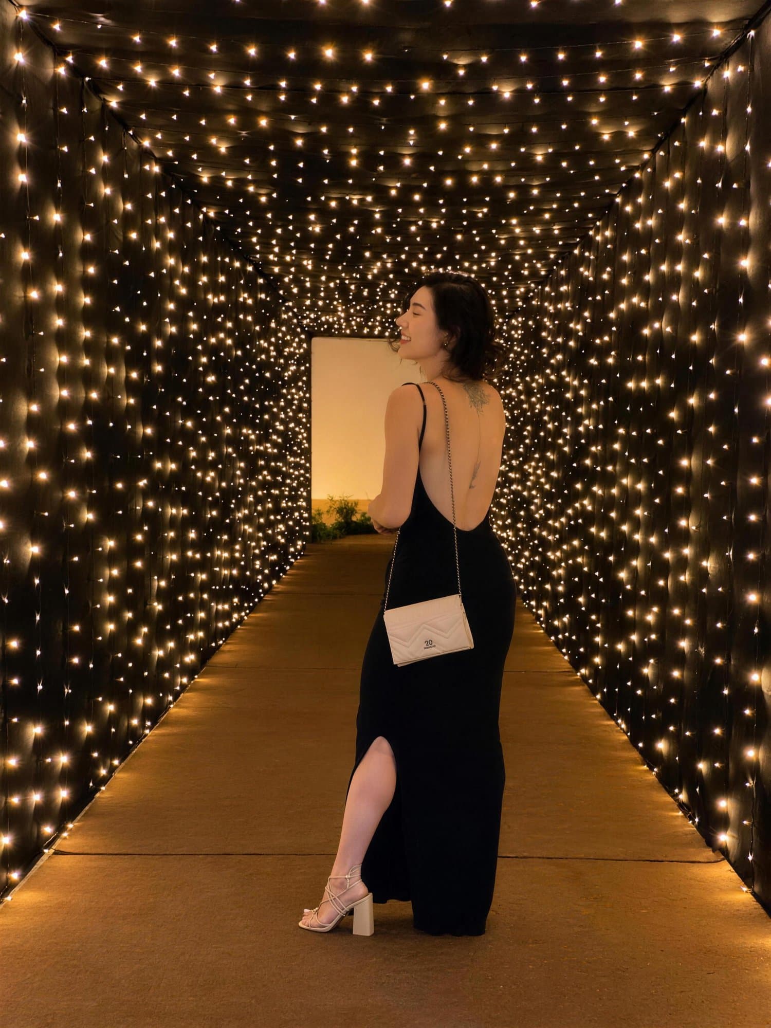 Camila de vestido preto, de costas, olha para trás num túnel de luzes.