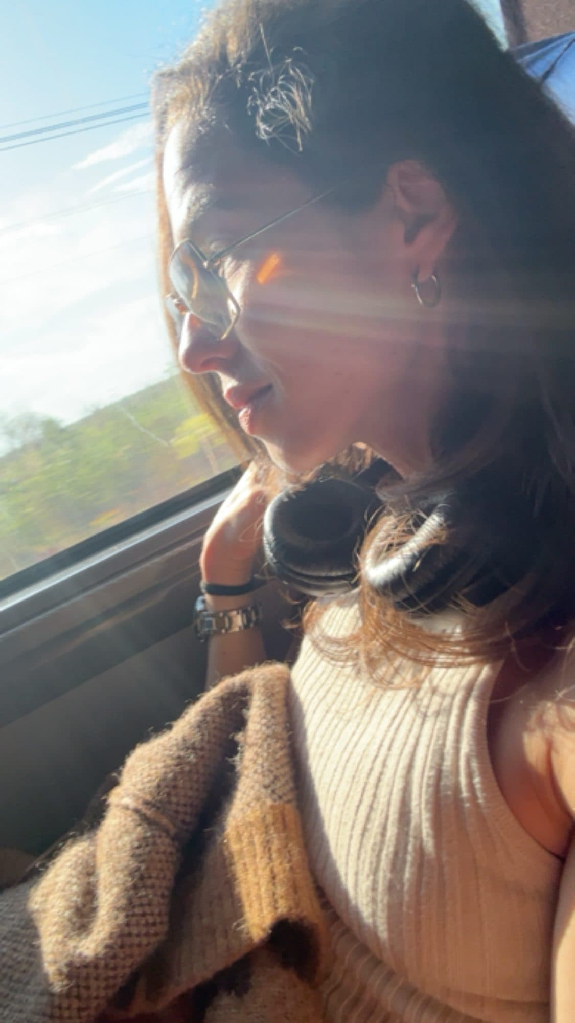 Camila sentada junto à janela do ônibus, com fones no pescoço e luz do sol no rosto.