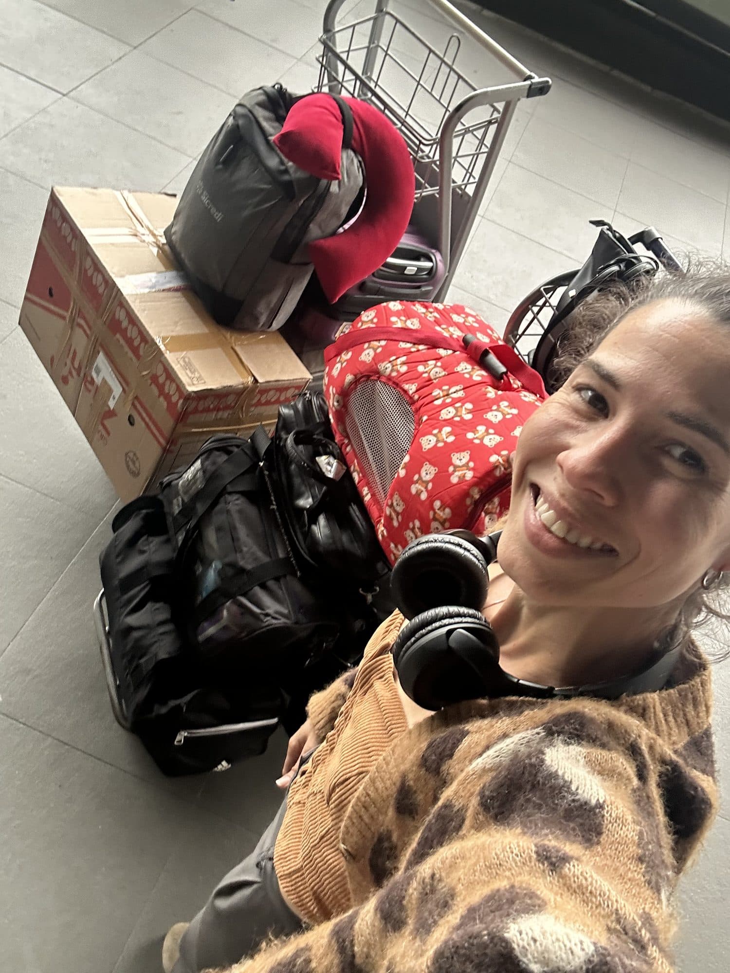 Camila sorri sentada no chão ao lado de malas, caixas e da caixa de transporte dos gatos.