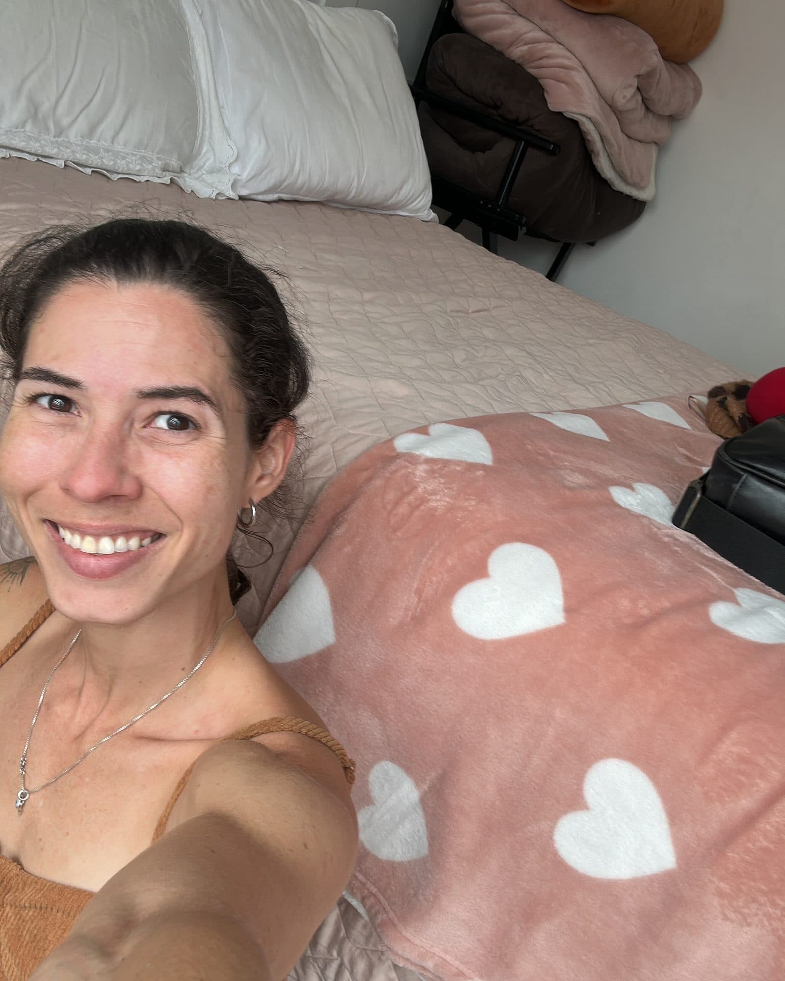 Camila sorri deitada na cama, ao lado de uma manta rosa com corações.